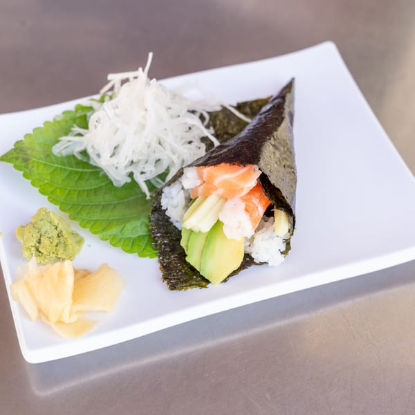 A salmon and avocado hand roll with rice, seaweed, wasabi, ginger, and daikon radish on a white plate.