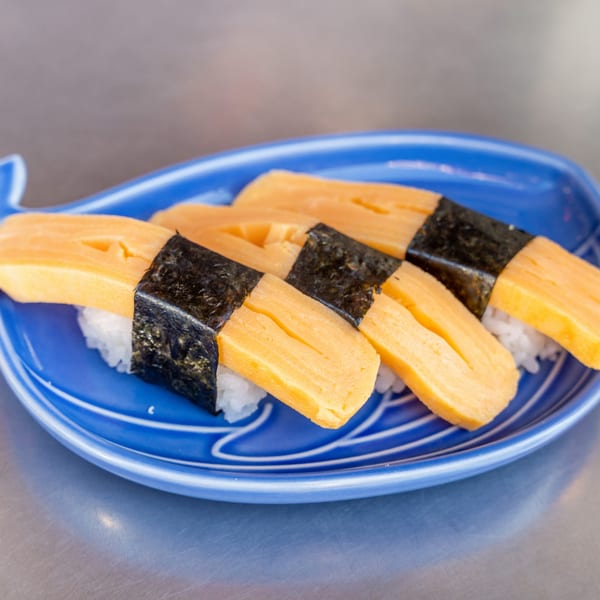 Four pieces of tamago nigiri sushi, with egg omelet and nori, on a blue whale-shaped plate.