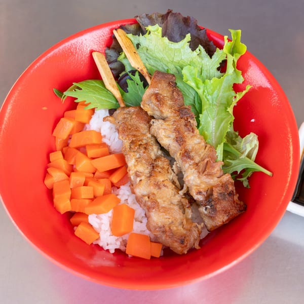 Two grilled meat skewers with rice, diced carrots, and fresh greens in a red bowl.