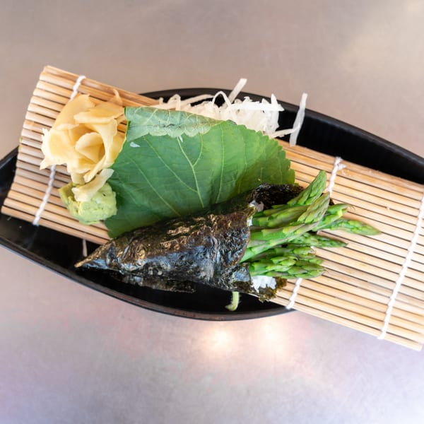 Asparagus tempura hand roll with ginger, wasabi, and a green leaf on a bamboo mat.