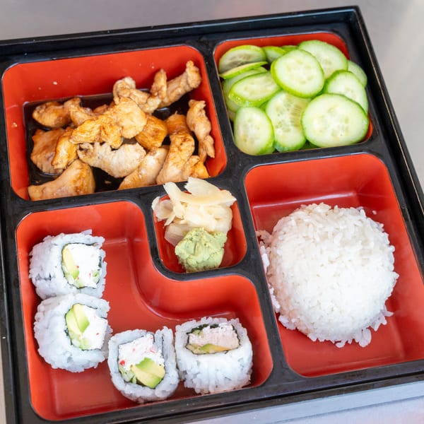 A bento box with teriyaki chicken, sliced cucumbers, sushi rolls, rice, ginger, and wasabi.